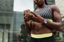 Ernte Afroamerikanerin in Sportbekleidung surft auf modernem Smartphone, während sie während des Trainings im Freien auf verschwommenem Hintergrund der Stadtstraße steht — Stockfoto