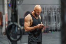 Seitenansicht eines muskulösen, glatzköpfigen Sportlers, der in einer modernen Turnhalle steht und während der Trainingspause auf dem Smartphone surft — Stockfoto