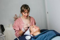 Cosmetólogo aplicando mascarilla facial hidratante en la cara del cliente femenino durante el procedimiento de cuidado de la piel en el salón de belleza moderno - foto de stock