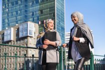 Lächelnde muslimische Unternehmerinnen mit Koffer spazieren durch die Stadt und schauen einander an — Stockfoto