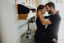 Homem barbudo alegre abraçando mulher sorridente enquanto se inclina no armário com pia na cozinha acolhedora em casa — Fotografia de Stock