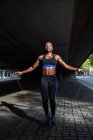 Femme afro-américaine en tenue de sport tenant la corde à sauter et regardant la caméra tout en se tenant sur le trottoir sur la rue de la ville — Photo de stock