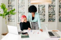Jovens designers de moda feminina multirracial discutindo esboços para nova coleção enquanto trabalham juntos na mesa na sala de luz — Fotografia de Stock