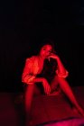 Sensuelle jeune hispanique ethnique femelle en lingerie regardant la caméra tout en se reposant sur la terrasse sous néon rouge — Photo de stock