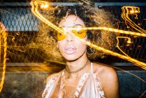 Femme afro-américaine auto-assurée avec des cheveux bouclés dans des lunettes de soleil à la mode et haut relaxant dans la rue le soir près de lumières gelées — Photo de stock