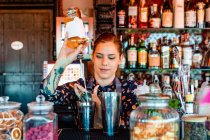 Garçonete feminina derramando álcool em shaker enquanto prepara coquetel refrescante no balcão no bar e olhando para longe — Fotografia de Stock