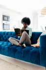 Junge schwarze Frau in lässigem Outfit mit Kopfhörern sitzt auf gemütlichem blauem Sofa zu Hause und liest Magazin, während sie die Freizeit zu Hause genießt — Stockfoto