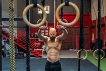 Сильний без сорочки чоловік дивиться вниз стоячи вправу на гімнастичних кільцях під час інтенсивного тренування в сучасному тренажерному залі — стокове фото