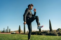 Взрослый спортсмен в спортивной одежде поднимает ногу и смотрит вперед во время тренировки на газоне под солнечным светом — стоковое фото