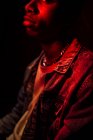 Cultiver calme homme afro-américain élégant en veste de jeans sous néon lumière rouge à l'ombre sur fond noir détournant les yeux — Photo de stock