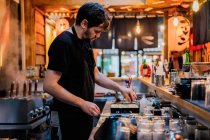 Vista lateral del joven en delantal cocinando platos asiáticos mientras está de pie en el mostrador en el bar ramen - foto de stock