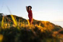 Снизу привлекательная молодая женщина в красном сарафане и шляпе стоит на зеленом травянистом лугу в солнечной сельской местности — стоковое фото