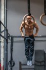 Full body shirtless bearded sportsman hanging on bar and doing pull ups during intense training in contemporary gym looking at camera — Stock Photo
