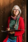 Artista donna anziana deliziata con tavolozza di colori e pennelli in piedi vicino alla porta di legno e guardando altrove — Foto stock