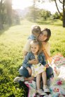 Jovem mãe feliz com filhinhas bonitos em roupas semelhantes desfrutando de dia ensolarado de verão juntos enquanto sentados em cobertor no gramado gramado gramado no parque — Fotografia de Stock