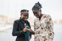 Trendige lächelnde Afroamerikanerinnen mit Frisur verbringen bei hellem Tag Zeit miteinander beim Handy-Surfen im Park — Stockfoto