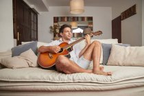 Дорослий чоловік у повсякденному одязі, сидячи на дивані, грає на акустичній гітарі у світлій вітальні та дивиться в сторону — стокове фото