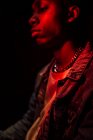 Crop calmo homem afro-americano elegante em jaqueta de jeans sob luz vermelha néon na sombra no fundo preto olhando para longe — Fotografia de Stock