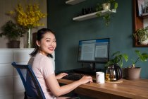 Vista lateral de la mujer freelancer asiática feliz leyendo documentos en el monitor de la computadora mientras está sentado mirando la cámara en la mesa durante el trabajo remoto - foto de stock