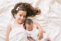 Top view of cheerful little girl embracing adorable infant while lying on soft bed at home — Stock Photo