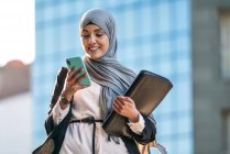 Empresária muçulmana feliz no hijab e com pasta em pé na rua navegando no telefone celular enquanto discute projeto de negócios — Fotografia de Stock