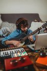 Молодий чоловік в навушниках грає на гітарі біля столу з ноутбуком і синтезатором вдома дивлячись на камеру — стокове фото