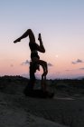 Vista lateral de una mujer flexible balanceándose boca abajo mientras practica acroyoga con su pareja masculina contra el cielo al atardecer en las montañas - foto de stock