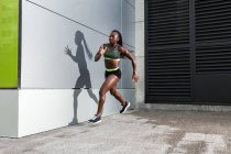 Бічний вид афроамериканської спортсменки стрибнув уперед, бігаючи біля сучасної будівлі на вулицях міста. — стокове фото