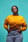 Mulher afro-americana rechonchuda sem emoção em camisola amarela olhando para a câmera com as mãos no bolso contra a parede azul — Fotografia de Stock