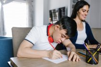 Этнический подросток с синдромом Дауна рисует карандашами на бумаге, сидя за столом с женщиной-фрилансером, работающей над ноутбуком дома — стоковое фото