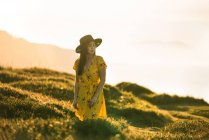 Attraente giovane femmina in giallo prendisole e cappello in piedi su verdeggiante prato erboso in campagna soleggiata — Foto stock