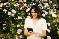 Fröhliche junge Frau meldet sich per Handy, während sie im Frühlingsgarten vor einem blühenden Strauch mit rosa Blüten steht — Stockfoto