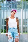 Queer male with bright pink hair and colorful nails standing in street and leaning on metal fence while looking at camera — Stock Photo