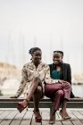 Élégant souriant à la mode afro-américaine dames passer du temps ensemble assis sur un banc bas en bois dans le parc dans une journée lumineuse en regardant la caméra — Photo de stock