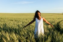 Усміхнена молода чорна леді в білій літній сукні, що ходить на зеленому пшеничному полі, дивлячись на камеру вдень під блакитним небом — стокове фото