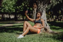 Mujer afroamericana en ropa deportiva sosteniendo un teléfono inteligente y tomando una selfie mientras está sentada en la hierba en un día soleado - foto de stock