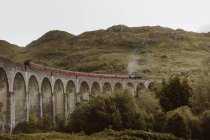 Steam train riding along old arch bridge near rough hill on gray day in Glenfinnan, UK countryside — Stock Photo