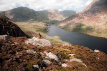 Back view of anonymous female admiring hills and river while sitting on rough slope during trip through Snowdonia, UK countryside — Stock Photo