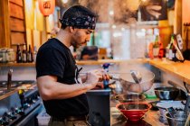 Vista lateral do homem em bandana de pé no balcão e cozinhar ramen no moderno café asiático — Fotografia de Stock