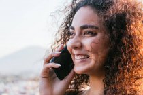 Zufriedene junge hispanische Frau mit langen lockigen Haaren, die mit dem Handy telefoniert und an einem sonnigen Sommerabend im Freien glücklich lächelt — Stockfoto