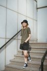 Jovem do sexo feminino em vestuário casual olhando para a câmera de pé em escadas contra parede de concreto do edifício moderno na rua urbana durante o dia — Fotografia de Stock