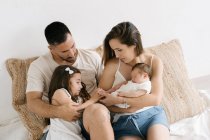 Cute little girl sitting on couch with smiling happy young parents holding newborn baby — Stock Photo