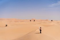 Lächelnde junge Frau in lässiger Kleidung, die während einer Reise in den Emiraten auf einer Sanddüne in der Wüste steht und die Arme ausstreckt — Stockfoto