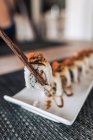Pessoa de colheita segurando com pauzinhos fileira de saborosos rolos de sushi com arroz cozido e fatias de frutos do mar em prato cerâmico na mesa — Fotografia de Stock