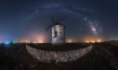 Malerische Landschaft der Milchstraße im dunklen Nachthimmel über einem alten steinernen Windmühlenturm mit leuchtenden Lichtern in der Ferne — Stockfoto