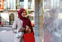 Compradora muçulmana encantada em lenço de cabeça e com sacos de compras de pé perto vitrine da loja na cidade — Fotografia de Stock