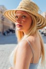 Vista lateral da encantadora fêmea usando chapéu de palha olhando para a câmera no dia ensolarado na rua da cidade no verão — Fotografia de Stock
