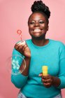 Fröhliche Afroamerikanerin mit geschlossenen Augen trägt blaue Kleidung und pustet Seifenblasen vor rosa Hintergrund im Studio — Stockfoto