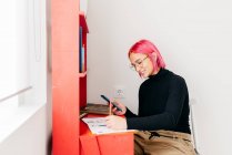 Vista lateral de la joven diseñadora creativa con pelo rosa en traje casual y gafas usando smartphone y boceto de dibujo mientras trabaja en el escritorio en casa - foto de stock