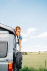 Full body black female tourist with curly hair looking up and climbing up ladder on modern RV against blue sky and green field in countryside — Stock Photo